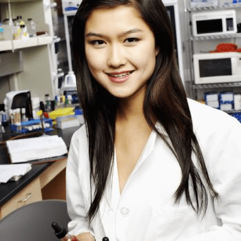 A woman wearing a white lab coat stands in a laboratory, holding lab equipment and smiling at the camera.