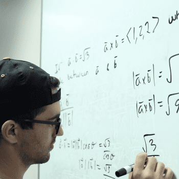 Person wearing a cap and glasses writes mathematical equations involving square roots and vectors on a whiteboard with a black marker.