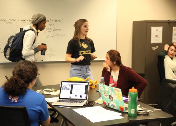 Four students are gathered in a classroom, some seated at a table with laptops and notebooks, while one stands and speaks; a whiteboard with notes is in the background.