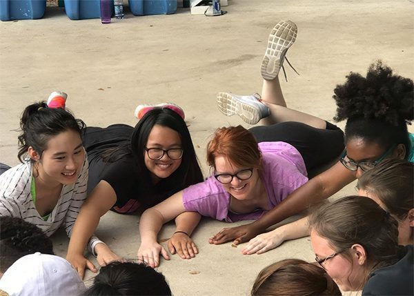 Four young women lying on the ground, smiling and reaching their hands toward a group in front of them, outdoors on a concrete surface.
