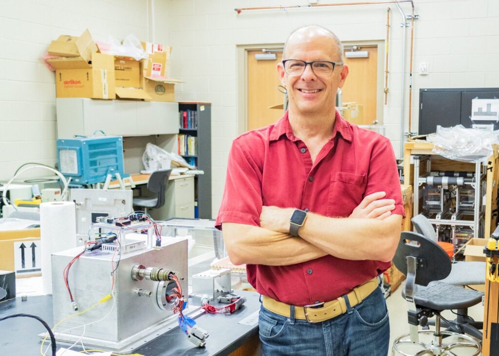 A person wearing glasses and a red shirt stands with arms crossed in a laboratory, surrounded by scientific equipment and electronics.