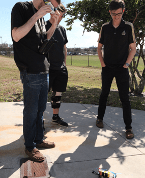 Three people stand outside on a sidewalk observing a small robotic device on the ground; one person is taking a photo or video.
