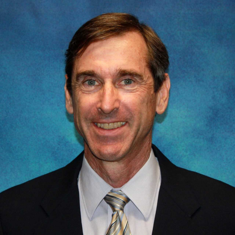 A middle-aged man in a suit and tie smiles at the camera against a blue textured background.