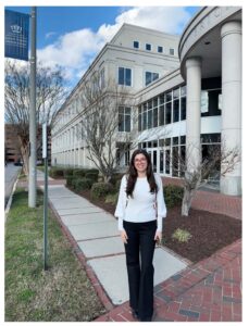 A woman wearing a white blouse and black pants stands smiling on a sidewalk in front of a large, modern building with columns and glass windows.