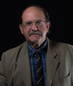A middle-aged man with glasses, a mustache, and thinning hair, wearing a tan blazer, plaid shirt, and striped tie, sits against a black background.