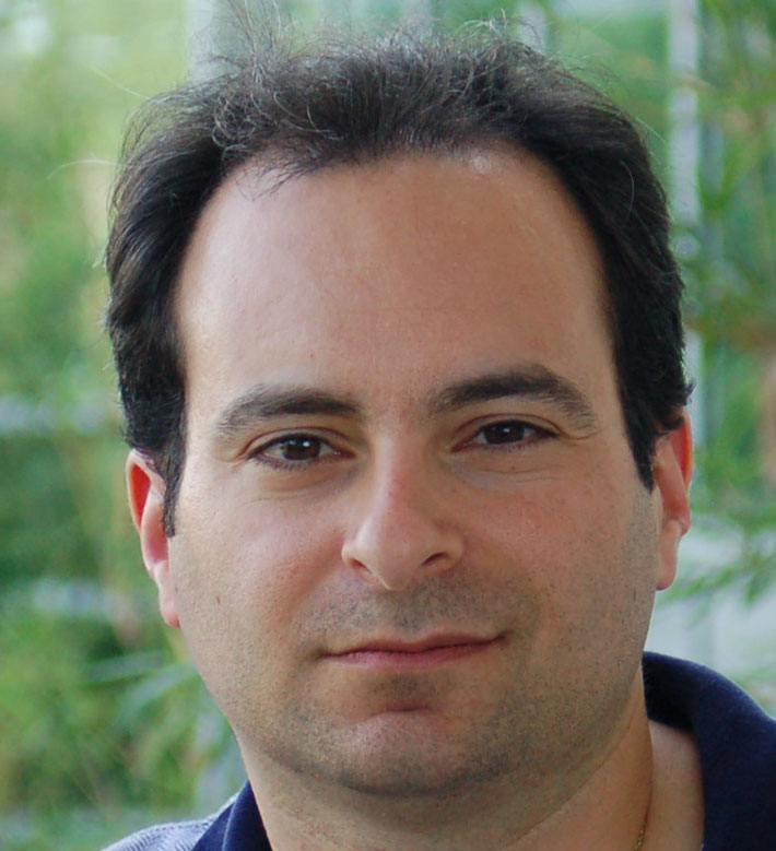 A man with short dark hair and fair skin looks directly at the camera, with a blurred green background behind him.