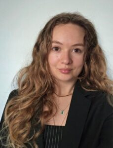 A young woman with long wavy hair wearing a black blazer and striped top, standing in front of a plain light background.