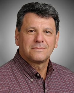 A middle-aged man with short dark hair wearing a maroon checked shirt poses against a neutral gray background.