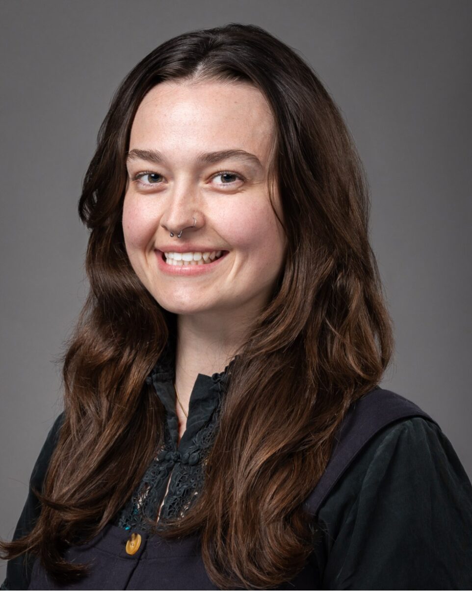 A person with long brown hair, wearing a dark blouse and vest, smiling at the camera against a plain gray background.