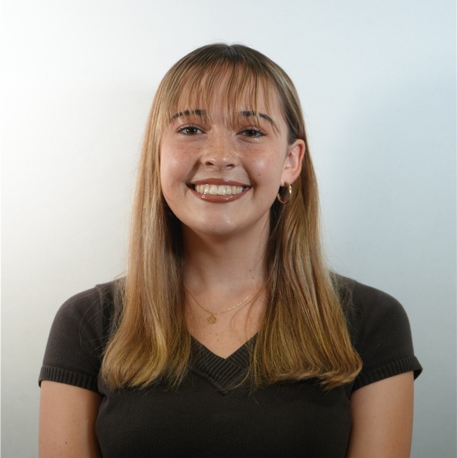 Young woman with straight blonde hair and bangs, wearing a dark short-sleeve top and gold jewelry, smiling in front of a plain white background.