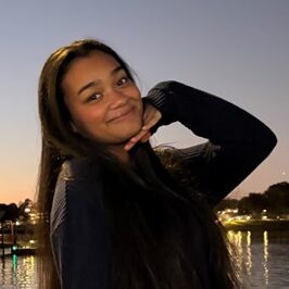A young woman with long dark hair poses by a waterfront at dusk, smiling with her hand under her chin. The sky is clear, and lights reflect on the water.