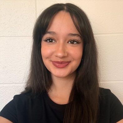 A young woman with long dark hair and a black shirt smiles at the camera, standing in front of a white brick wall.