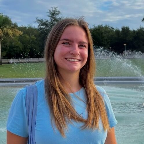 A young woman in a light blue shirt stands smiling in front of a water fountain with trees and a partly cloudy sky in the background.