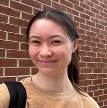 A person with long dark hair, wearing a tan shirt and black backpack, stands in front of a brick wall and smiles at the camera.