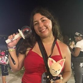 A woman in a red top smiles while holding a light stick and a plush doll at an outdoor event at night. Other people are visible in the background.