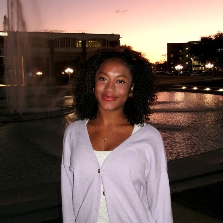 A woman with curly hair stands in front of a fountain at sunset, wearing a light purple cardigan and white top, smiling at the camera.