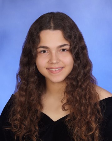 A young person with long, curly brown hair and a slight smile poses in front of a blue background, wearing an off-the-shoulder black top.