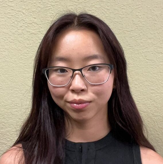 A young woman with long dark hair and glasses stands against a textured beige wall, wearing a sleeveless black top and looking at the camera.