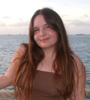 A young woman with long brown hair stands by a rope barrier in front of a body of water, wearing a brown top and denim shorts. The sky is partly cloudy at sunset.