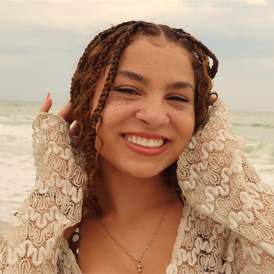 Young woman with braided hair smiling at the camera, standing on a beach with waves and cloudy sky in the background, wearing a patterned long-sleeve top.