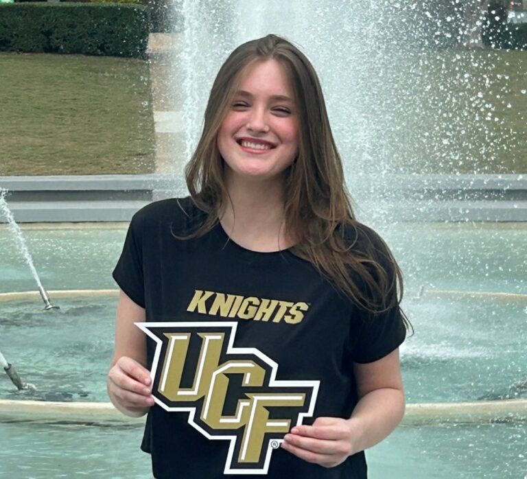Smiling person stands in front of a fountain, wearing a "Knights" shirt and holding a UCF sign.