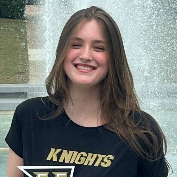 Smiling person stands in front of a fountain, wearing a "Knights" shirt and holding a UCF sign.
