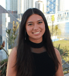 A woman in a black dress stands on a rooftop with city buildings, a river, and a bridge in the background, smiling at the camera.