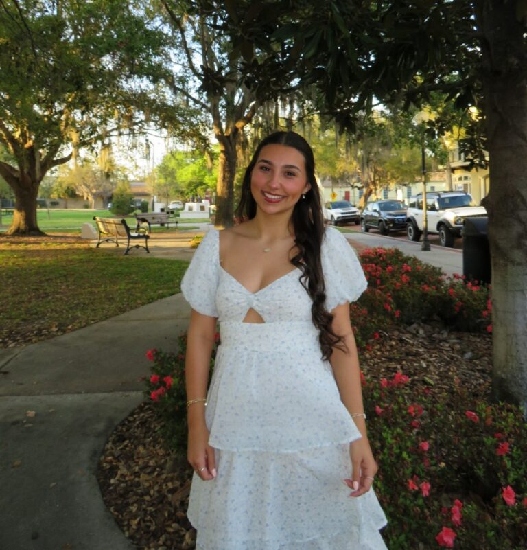 A woman in a white dress stands smiling in a park with trees, flowers, benches, and parked cars in the background.
