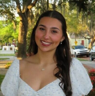 A woman in a white dress stands smiling in a park with trees, flowers, benches, and parked cars in the background.