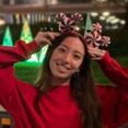 A woman in a red sweater and white pants poses at night with holiday lights and tree decorations in the background, wearing festive antler headbands.