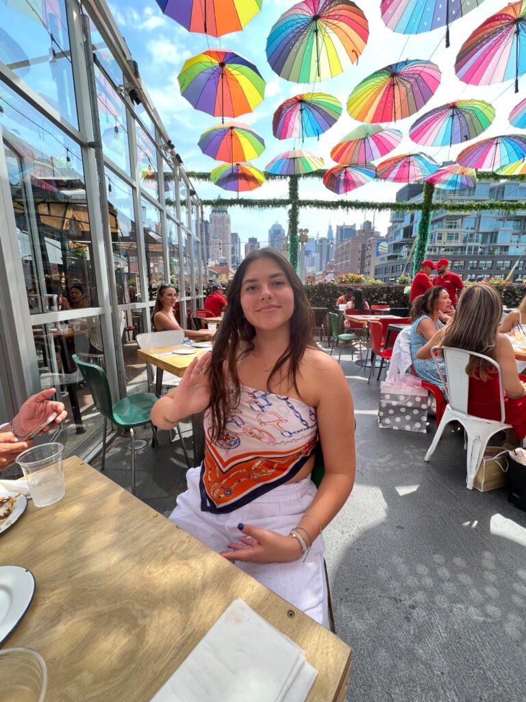 A woman sits at an outdoor restaurant table under colorful umbrella decorations, with other diners and a city skyline in the background.