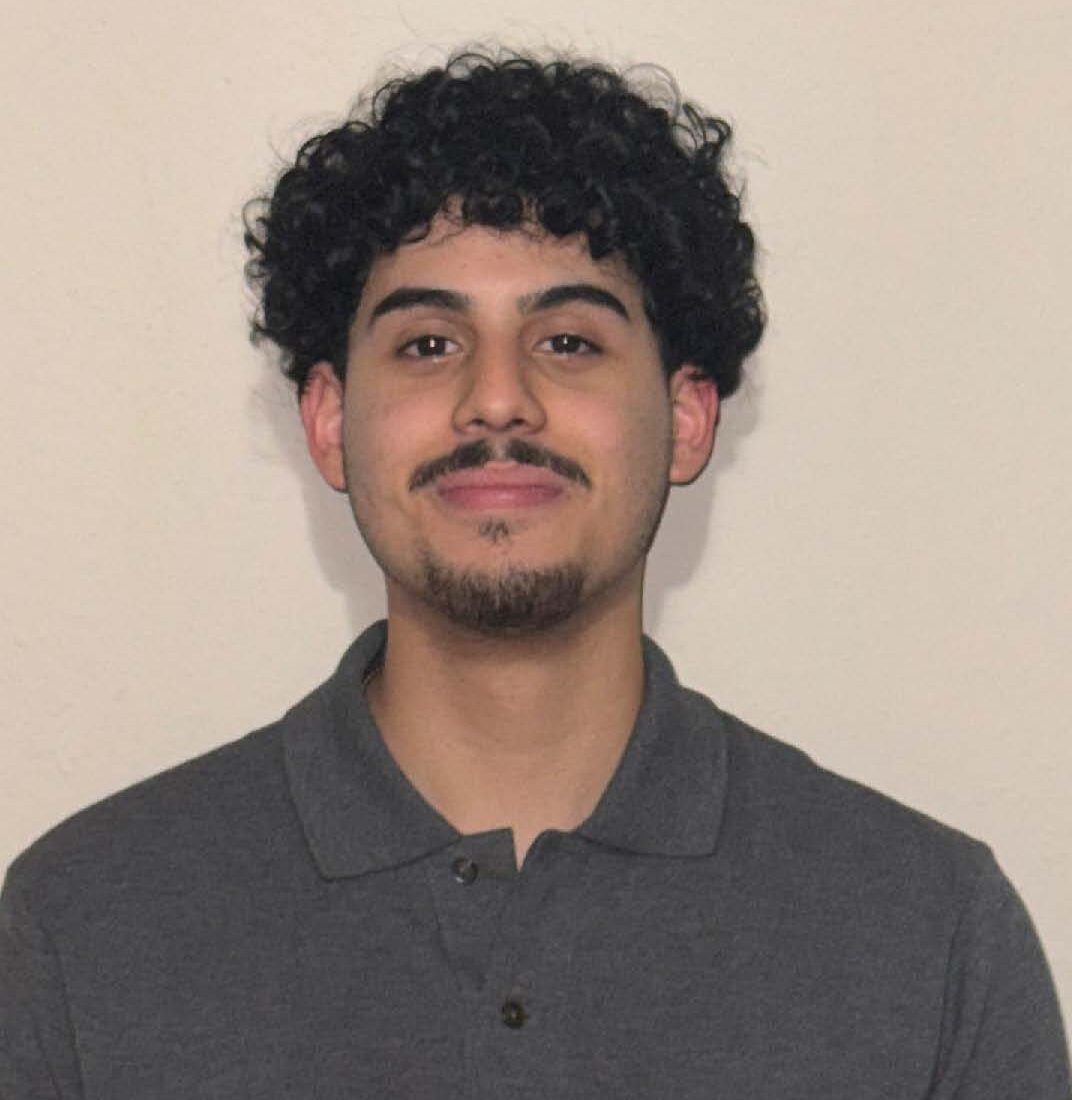 A young man with curly dark hair and a mustache is standing against a plain light-colored wall, wearing a dark grey polo shirt.
