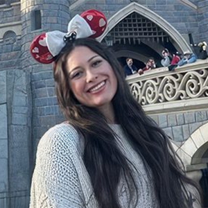 Woman with long dark hair wearing a white sweater and Minnie Mouse ears smiles in front of a castle-themed building at a theme park.