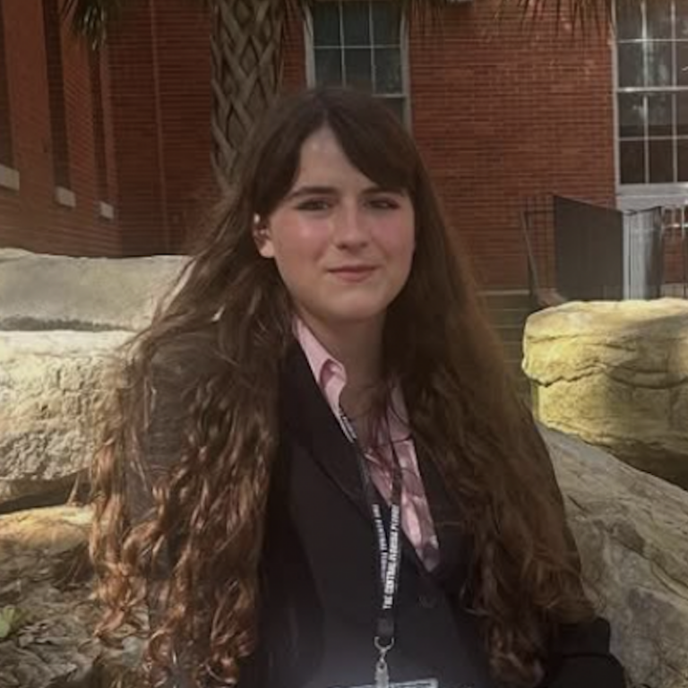 A person with long brown hair, wearing a black blazer and lanyard, sits outdoors on large rocks in front of a brick building.