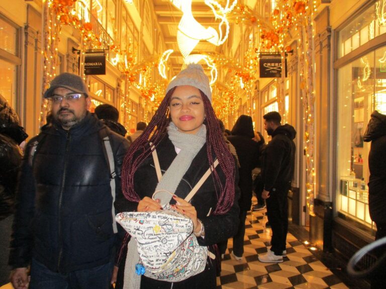 A woman in a gray hat and scarf stands indoors with festive golden lights and decorations overhead; people walk around her in a busy, well-lit corridor.