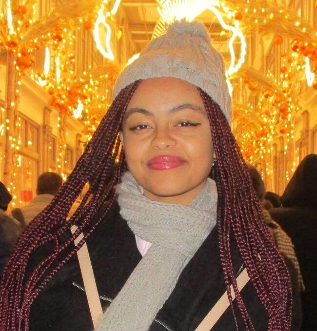 A woman in a gray hat and scarf stands indoors with festive golden lights and decorations overhead; people walk around her in a busy, well-lit corridor.