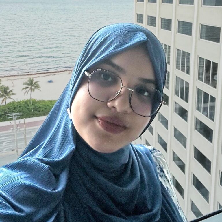 A woman in a blue hijab and glasses takes a selfie on a balcony with an ocean view, palm trees, and a tall building in the background.
