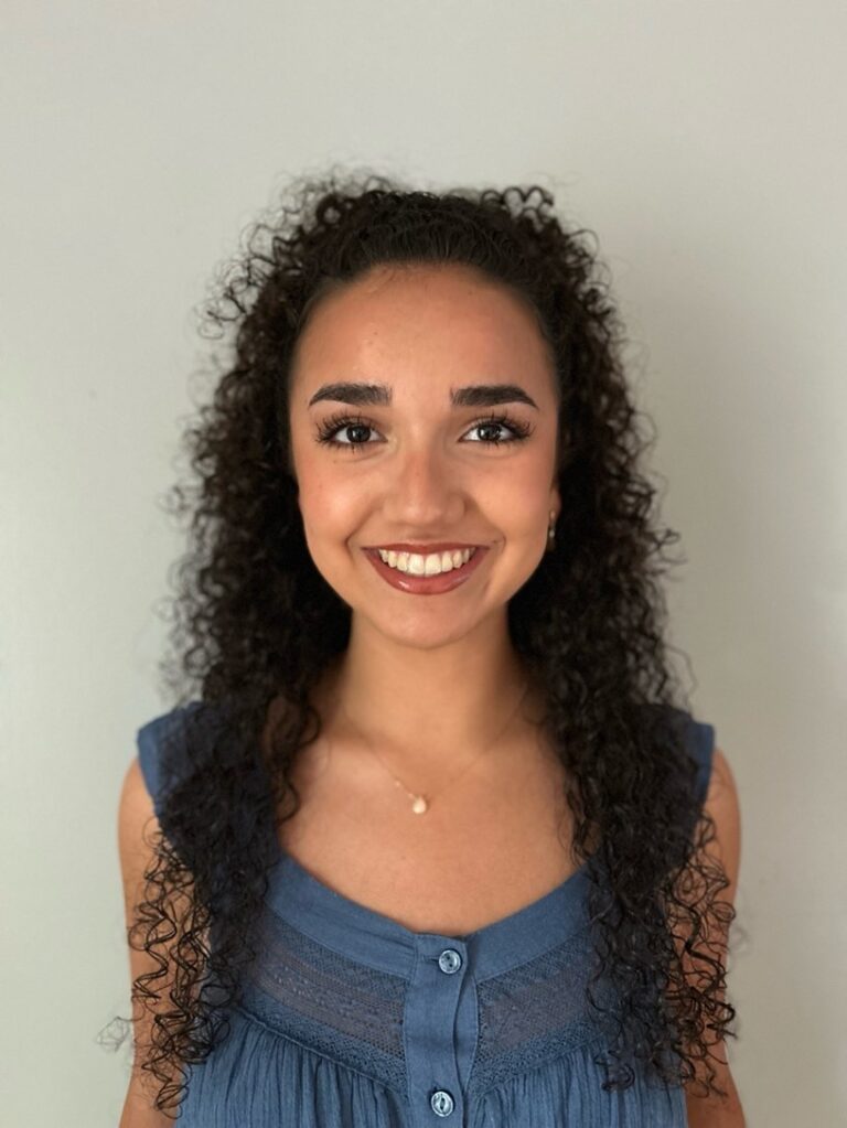 A young woman with curly dark hair, wearing a blue sleeveless top and a necklace, smiles at the camera against a plain light-colored background.