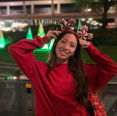 Woman in a red sweater poses outdoors at night, wearing a headband with peppermint candy decorations. Illuminated trees and blurred lights are in the background.