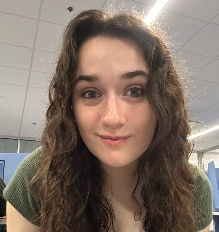 A person with long curly brown hair and a green top is leaning forward and looking at the camera in an office or classroom setting with blue chairs and overhead lighting.