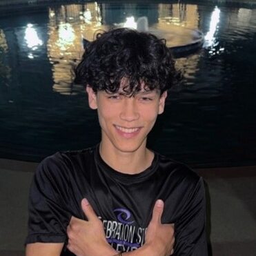 A young person stands poolside at night, smiling and crossing their arms in front of their chest, with a lit swimming pool in the background.