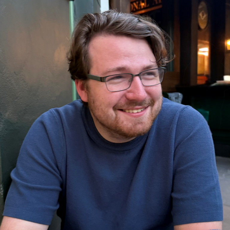 A man with glasses and a beard, wearing a blue short-sleeve shirt, sits and smiles outdoors near a green wall.