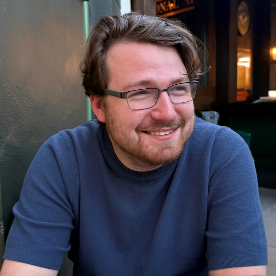 A man with glasses and a beard, wearing a blue short-sleeve shirt, sits and smiles outdoors near a green wall.