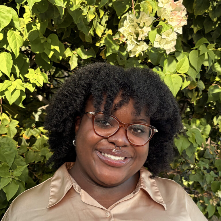 A person with curly hair and glasses smiles while standing in front of a leafy green background, wearing a light-colored shirt.