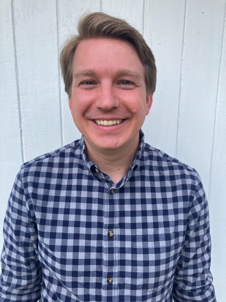 A person with short light brown hair, wearing a blue and white plaid button-up shirt, smiles while standing in front of a white wooden wall.