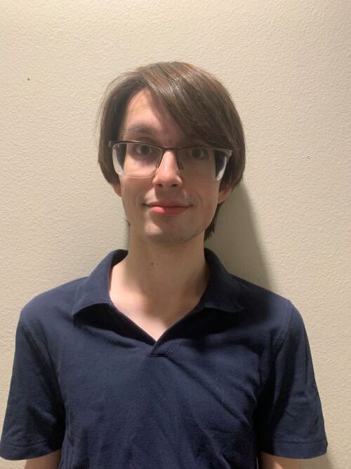 Young person with glasses and straight brown hair stands against a plain off-white wall, wearing a dark navy collared shirt and looking at the camera with a neutral expression.