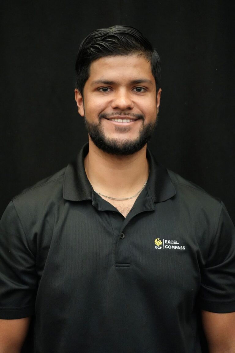 A man with short dark hair and a beard, wearing a black polo shirt with a "UCF Excel Compass" logo, stands in front of a plain black background and smiles at the camera.