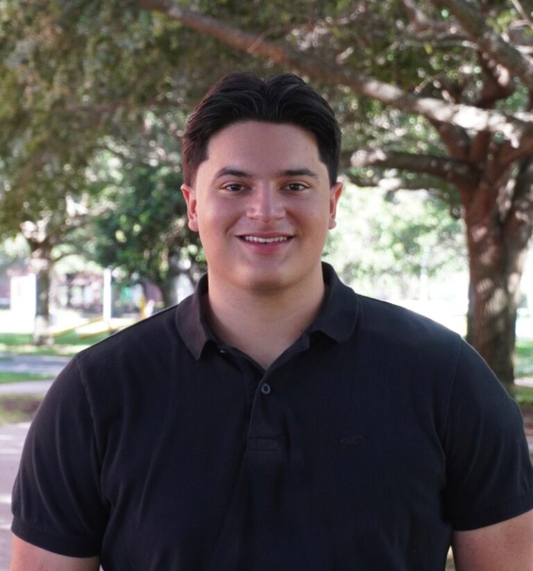 A man with dark hair in a black polo shirt stands outside, smiling, with leafy trees and sunlight in the background.