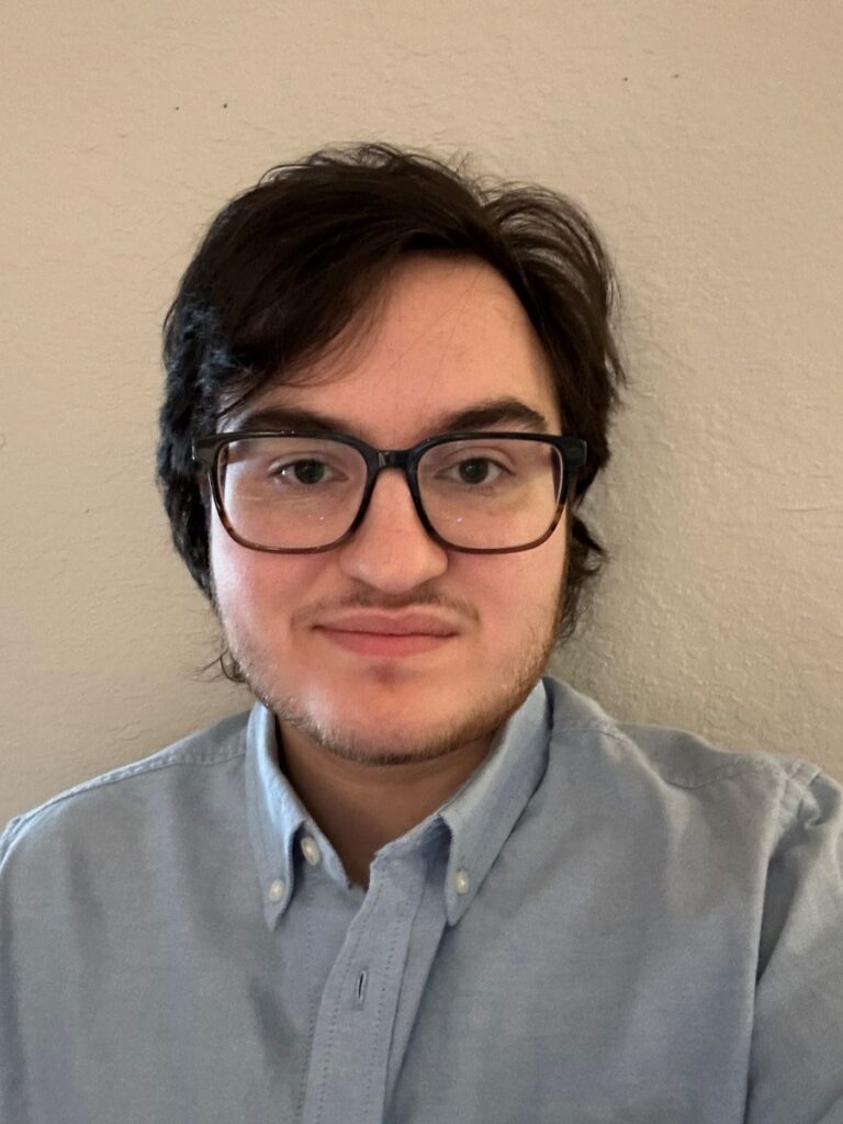 A person with short dark hair and glasses is wearing a light blue button-up shirt, standing in front of a plain beige wall.