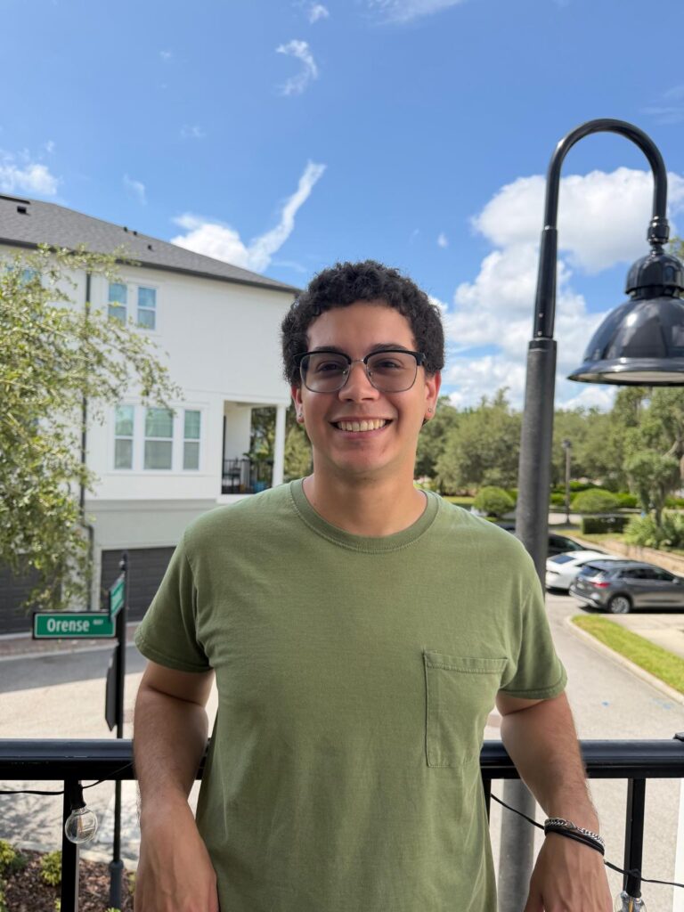A person wearing glasses and a green shirt stands on a balcony with trees, cars, and buildings visible in the background under a blue sky.